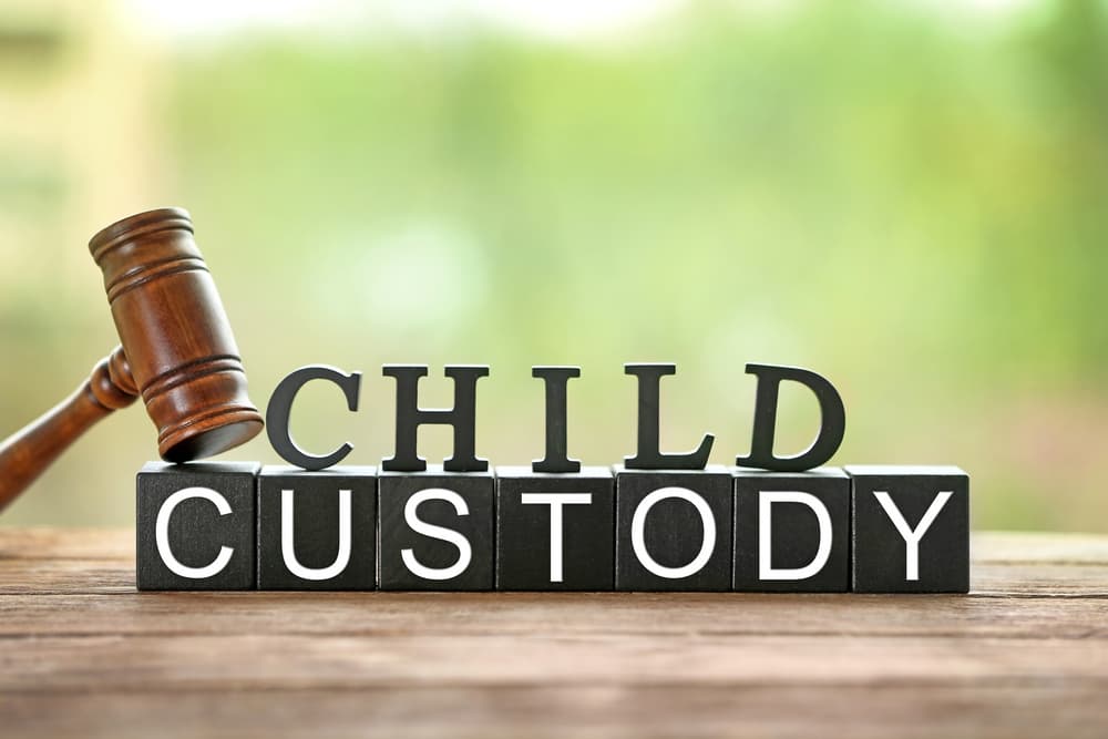 Text "CHILD CUSTODY" formed by black letter blocks with a judge's gavel on a table in the foreground, blurred background, close-up view.