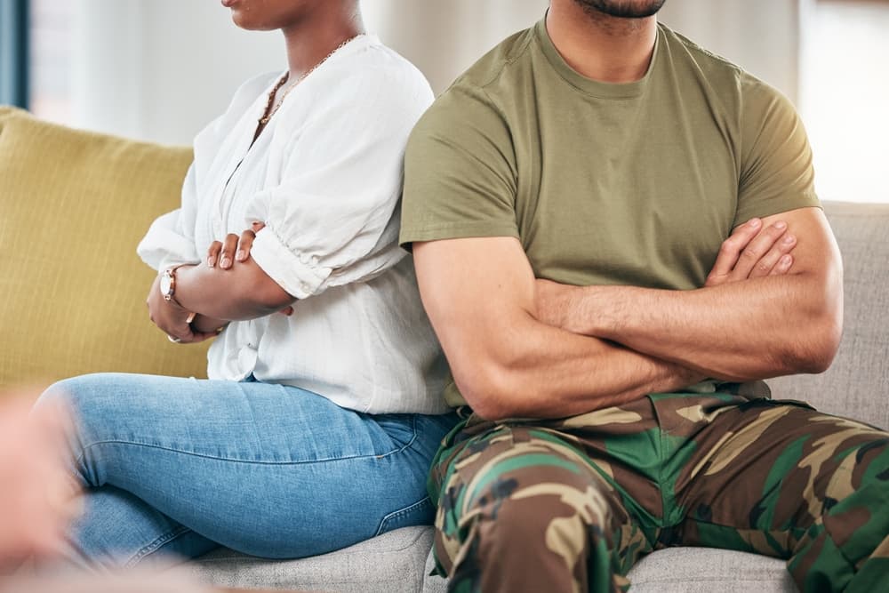 A tense couple sits on a sofa in their living room, arms crossed in frustration. The interracial couple, a man and a Black woman, faces relationship issues, their expressions and body language showing anger and conflict.