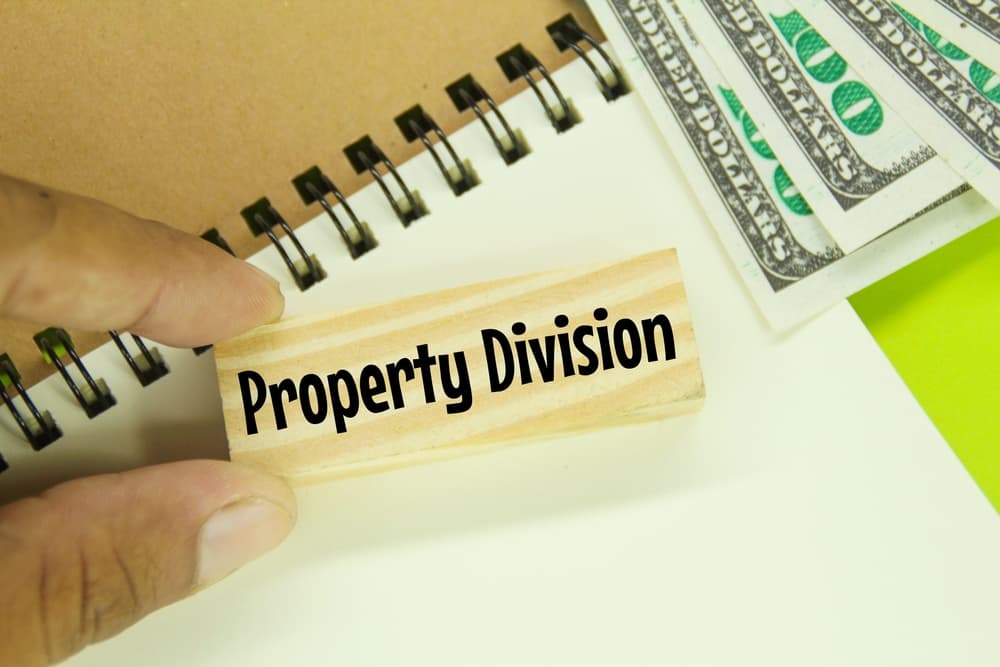 A stack of banknotes sits next to open notebooks on a wooden surface, with a rustic wooden board displaying the words "Property Division" etched or written on it. 